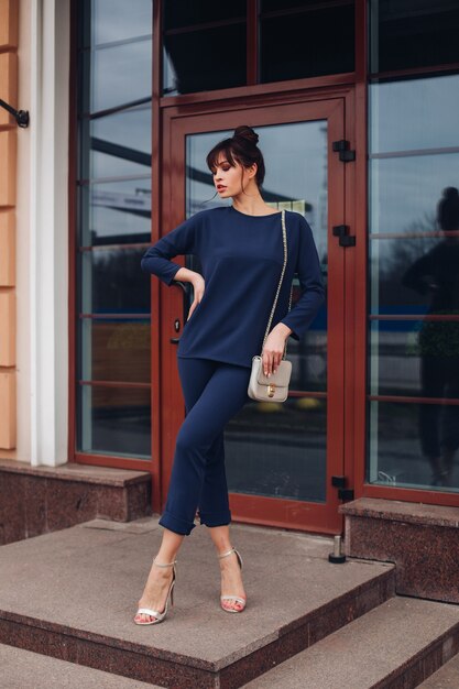 Photo d'une belle jeune femme caucasienne aux cheveux noirs en blouse bleu foncé et pantalon bleu foncé, chaussures blanches avec sac blanc