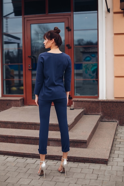 Photo d'une belle jeune femme caucasienne aux cheveux noirs en chemisier bleu foncé et pantalon bleu foncé, chaussures blanches avec sac blanc dos à la caméra