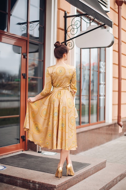 Photo gratuite la photo d'une belle jeune femme caucasienne aux cheveux noirs en robe dorée et aux chaussures dorées montre différents stands près du magnifique bâtiment