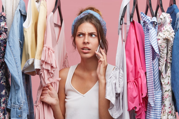 Photo de la belle jeune femme caucasienne inquiète debout à la garde-robe parmi les vêtements d'été et parler au téléphone portable à un ami lui demandant de l'aider tout en choisissant la robe parfaite à porter à la date