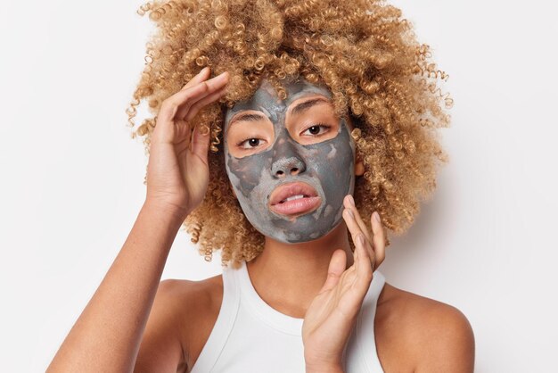 La photo d'une charmante femme bouclée sérieuse touche le visage applique doucement un masque facial en argile pour être belle porte un t-shirt décontracté pose sur fond blanc montre des épaules nues Concept de soins de la peau