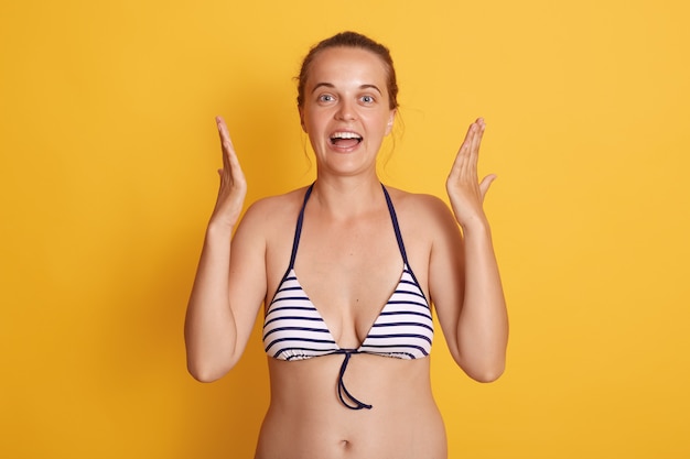 Photo de charmante jeune femme en maillot de bain rayé et montrant la taille avec les mains isolées sur le mur jaune, a l'air excité.