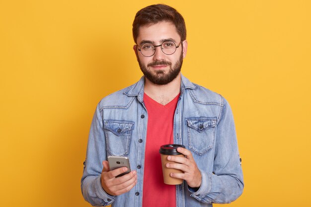 Photo de confiant beau jeune homme tenant une tasse de papier avec du café et un smartphone, portant une veste en jean, des lunettes et un t-shirt