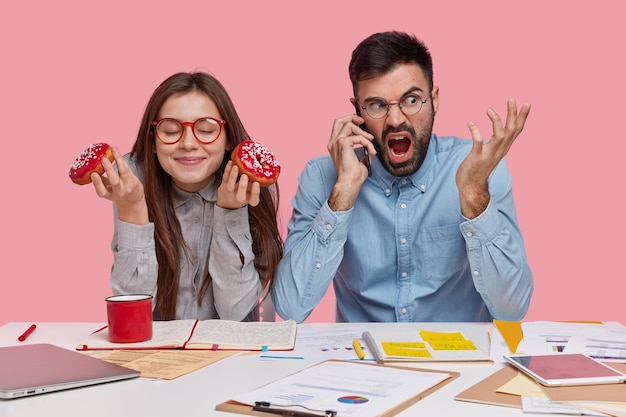 Photo d'une dame heureuse porte des lunettes à jante rouge, mange de délicieux beignets, s'assoit près de son partenaire masculin qui parle au téléphone intelligent avec une expression de colère