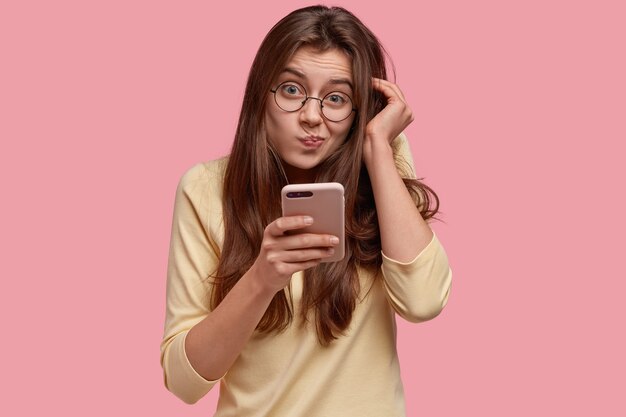 Photo d'une dame mécontente qui porte les lèvres et regarde avec une expression désemparée, tient un téléphone portable, surfe sur Internet, regarde l'écran, lit des nouvelles perplexes