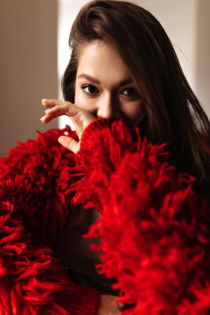 Photo d'une dame en veste rouge couvrant son visage avec sa main. Femme aux yeux bruns regardant la caméra.