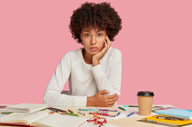 Photo d'un designer noir attrayant avec coupe de cheveux afro, assis sur le lieu de travail, porte un pull blanc, fait des dessins avec des crayons de couleur, isolé sur un mur rose, apprécie le café