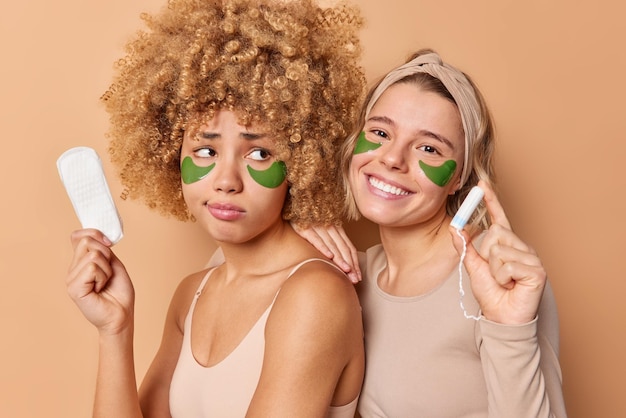 Photo de deux amies posent avec des produits d'hygiène choisir entre une serviette hygiénique et un tampon appliquer des patchs d'hydrogel sous les yeux subir des procédures de beauté se tenir étroitement l'une à l'autre à l'intérieur