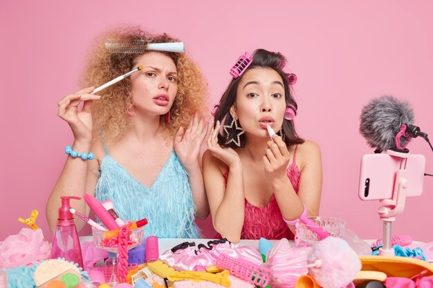 Photo de deux femmes blogueuses de beauté enregistrent une vidéo d'examen cosmétique pour un blog appliquent de la poudre et du rouge à lèvres se préparent pour la date font des coiffures frisées portent des robes à la mode posent à l'intérieur. La diffusion en direct