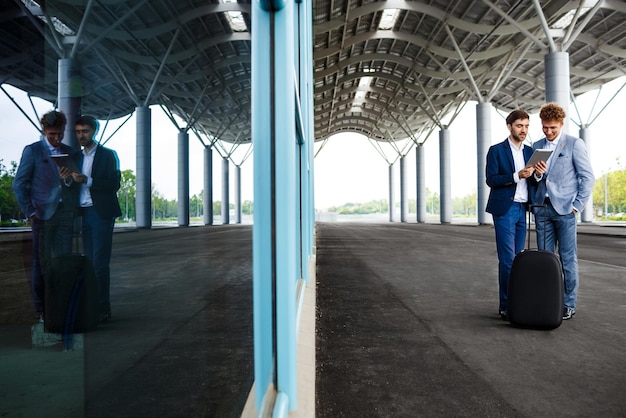 Photo gratuite photo de deux jeunes hommes d'affaires parlant sur fond de station et tenant une tablette reflétée dans la fenêtre