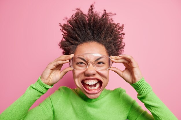 La photo d'une drôle de fille frisée folle a ouvert la bouche montre que les dents gardent les mains sur des lunettes transparentes porte un col roulé vert étant très émotif fait la grimace,