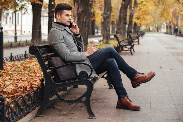 Photo de l'élégant homme brune en manteau et jeans boire du café à emporter et parler sur smartphone, assis sur un banc dans le parc