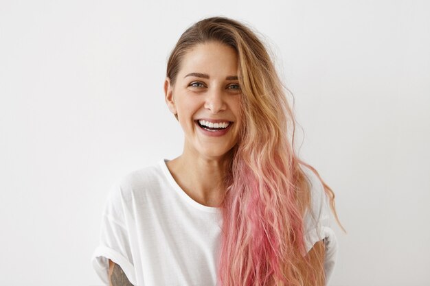 Photo de l'élégante jeune femme à la mode d'apparence européenne portant ses cheveux rose pastel en désordre sur le côté souriant largement, démontrant ses dents blanches parfaites. Jeunesse et bonheur