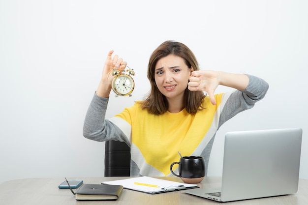 Photo d'une employée de bureau montrant le pouce vers le bas et tenant un réveil. Photo de haute qualité