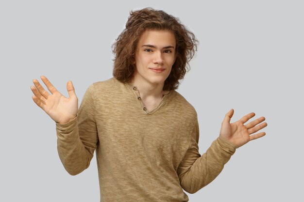 Photo d'un étudiant mignon avec des cheveux ondulés montrant des paumes ouvertes surélevées, démontrant ses bonnes intentions honnêtes. Séduisante jeune homme levant les mains, étant confus, posant