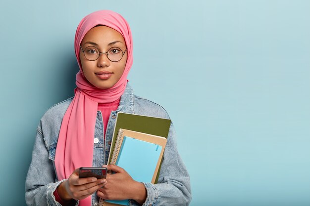 La photo d'un étudiant musulman porte un bloc-notes pour les notes, tient un cellulaire moderne, crée une nouvelle publication dans les réseaux sociaux, couvre la tête avec un voile selon les règles religieuses, discute en ligne avec ses camarades