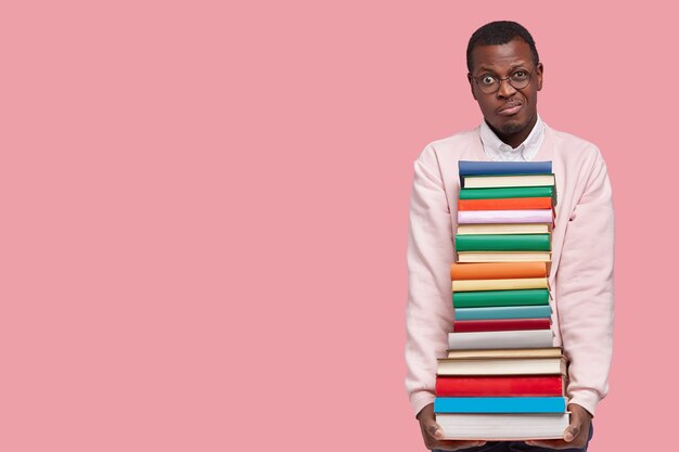 Photo d'un étudiant noir mécontentement tient une lourde pile de manuels, regarde avec mécontentement, se prépare à l'examen