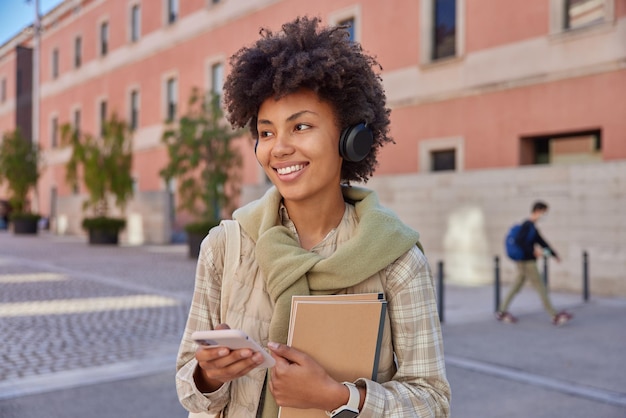 Photo gratuite photo d'une étudiante joyeuse écoute des informations via un casque stéréo utilise un smartphone porte des blocs-notes regarde ailleurs se promène joyeusement à l'extérieur sourit joyeusement concept de personnes et de style de vie