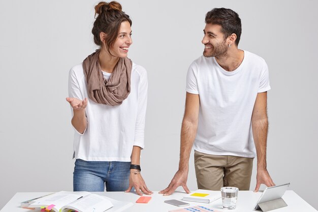 Photo d'experts en marketing à succès se réunissent pour discuter des principaux problèmes de travail, porter des vêtements à la mode, se regarder joyeusement, modèle près du bureau avec les choses nécessaires, isolé sur blanc