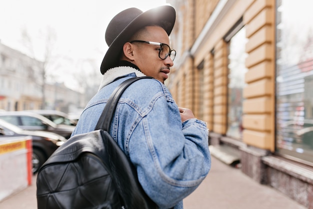 Photo extérieure de l'arrière d'un homme africain confiant en veste en jean marchant dans la rue. Élégant mec noir regardant la vitrine.