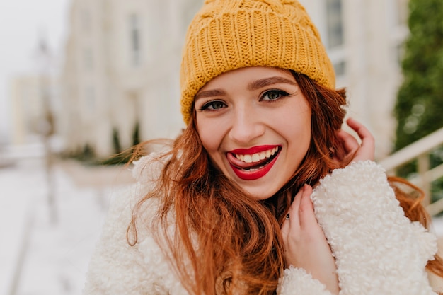 Photo extérieure d'une femme au gingembre ludique profitant de l'hiver. Rire jolie dame au chapeau jaune marchant en plein air.