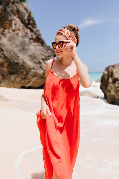 Photo extérieure de jolie fille blanche en longue robe rouge se promenant sur la plage. Un modèle féminin enthousiaste porte un ruban élégant posant avec des roches.