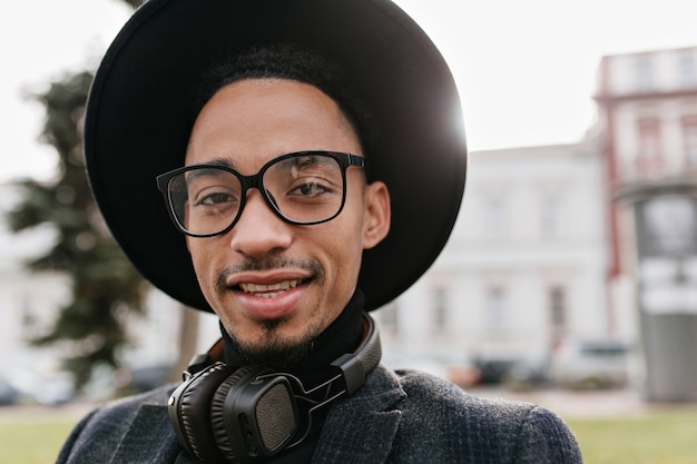 Photo extérieure de souriant jeune homme africain en grand chapeau posant. Portrait en gros plan d'un mec effrayant dans un casque noir, passer du temps dans le parc