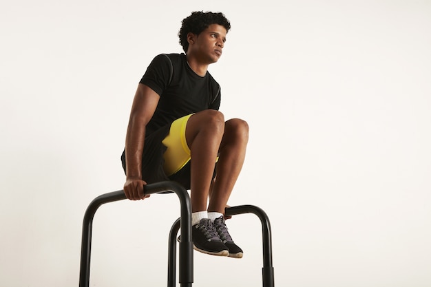 Photo à faible angle d'un modèle masculin noir maigre musclé fort avec un afro dans des vêtements d'entraînement noirs, levant les genoux sur des barres parallèles isolés sur blanc.