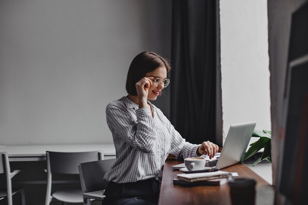 Photo de femme d'affaires aux cheveux courts à lunettes et chemisier blanc assis sur le lieu de travail et travaillant dans un ordinateur portable.