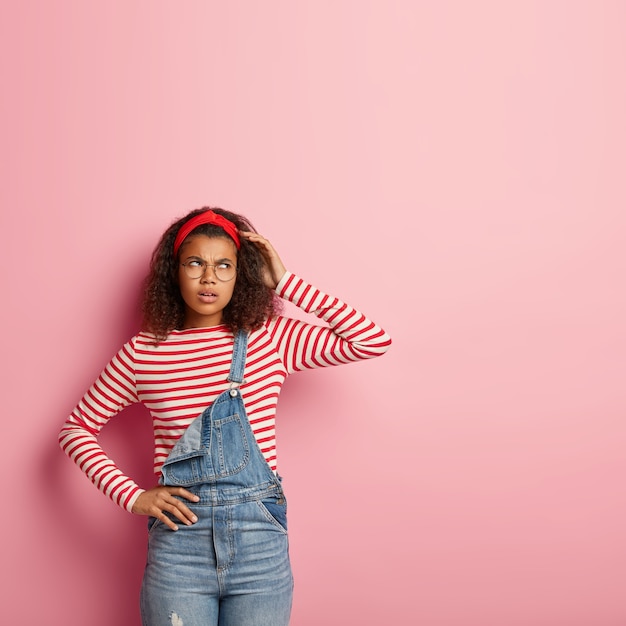 Photo D'une Femme Afro-américaine En Colère Réfléchie Avec Une Expression Pensive, Se Gratte La Tête, Porte Un Bandeau Rouge, Un Pull Rayé Et Une Salopette En Denim