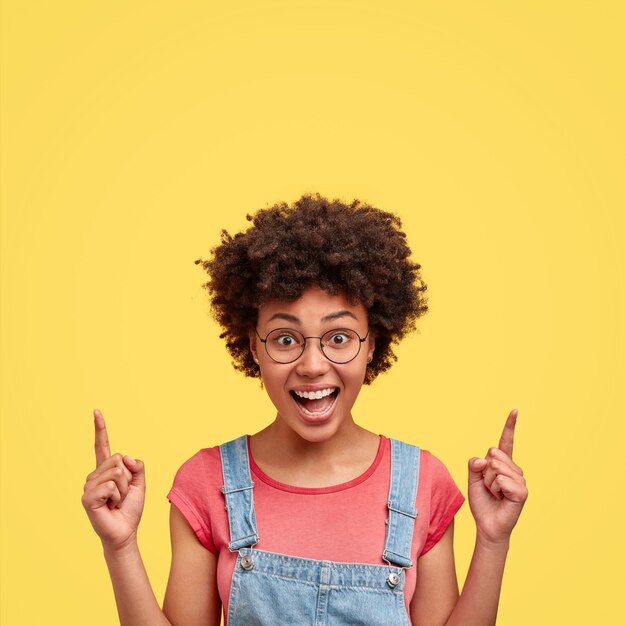 Photo d'une femme afro-américaine joyeuse avec un sourire positif, des cheveux nets, des points au-dessus avec l'index, a une expression joyeuse, pose contre le mur jaune. Heureuse femme à la peau sombre à l'intérieur