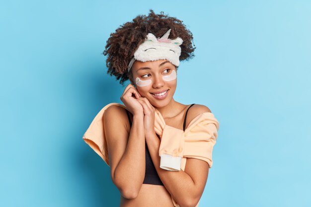 Photo gratuite photo d'une femme afro-américaine magnétique rêveuse concentrée de côté avec un sourire doux garde les mains près du visage applique des tampons de collagène sous les yeux