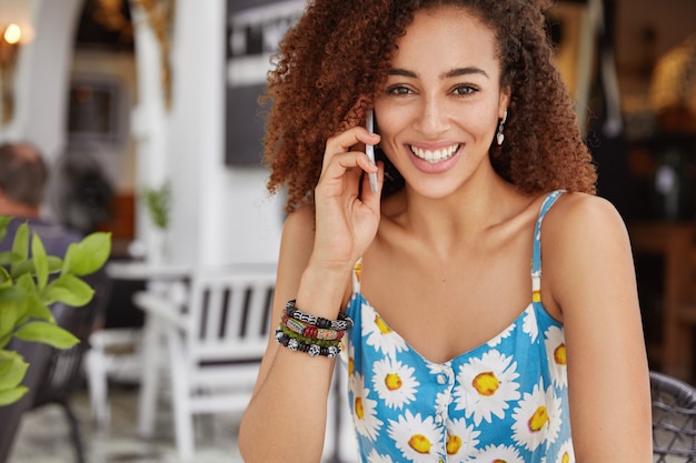 Photo de femme afro-américaine à la recherche agréable avec une expression joyeuse, appelle un ami par téléphone portable, bénéficie de temps libre pendant la journée d'été