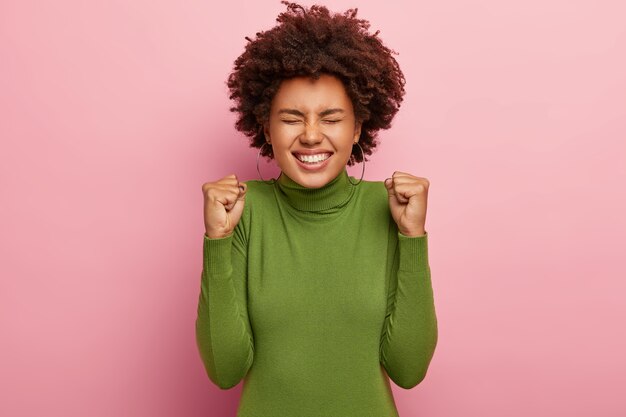 Photo d'une femme afro ravie, serre les poings avec triomphe, sourit largement, se réjouit de sa nouvelle réalisation