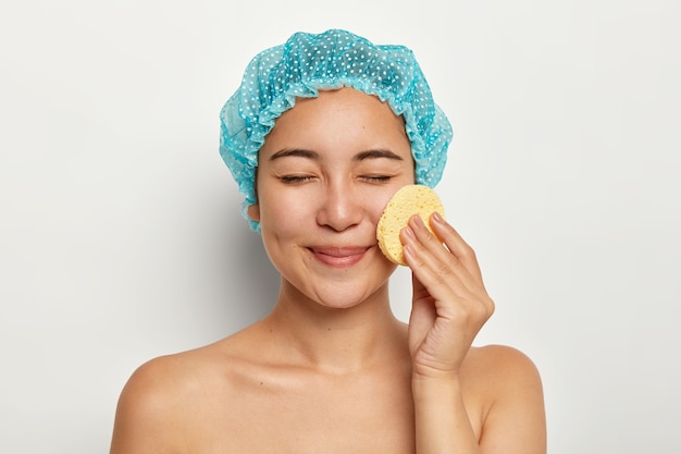 Photo d'une femme asiatique nettoie le visage avec une éponge de cellulose, se soucie de la peau, a des traitements de cosmétologie, les yeux fermés, se tient avec le corps nu, se tient contre un mur blanc. Procédures de spa, démaquillage