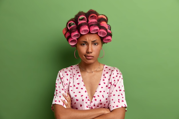 Photo de femme au foyer en colère sérieuse se tient avec les bras croisés sur le corps et l'expression mécontente, attend des explications du mari, porte un pyjama et des bigoudis, isolé sur vert