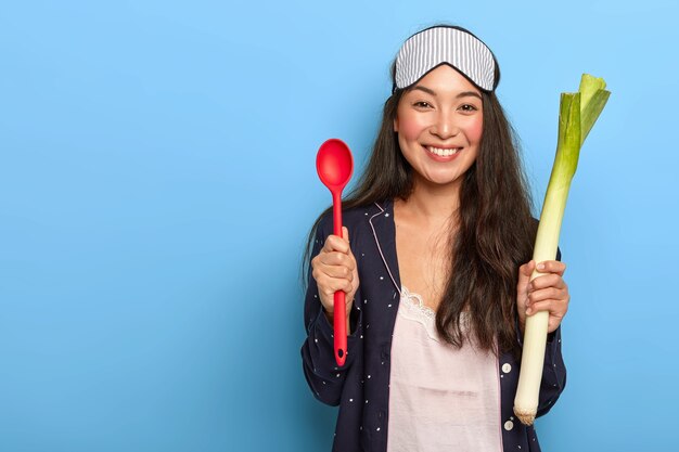 Photo de femme au foyer heureuse va cuisiner un plat sain le matin, tient le poireau vert et une cuillère rouge, sourit agréablement, porte des vêtements de nuit