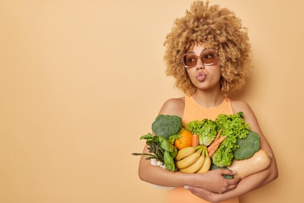 Photo d'une femme aux cheveux bouclés porte un tas de fruits et légumes frais garde les lèvres pliées porte des lunettes de soleil à la mode et un t-shirt décontracté isolé sur fond marron espace de copie vierge pour la promotion