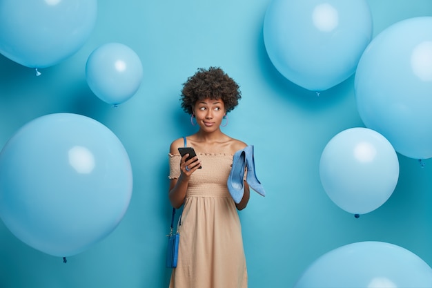 Photo d'une femme aux cheveux bouclés songeuse tient une paire de chaussures à talons hauts bleus et un téléphone portable, fait des achats en ligne, achète une tenue à la mode, isolée sur un mur bleu. Habillage, concept de vêtements