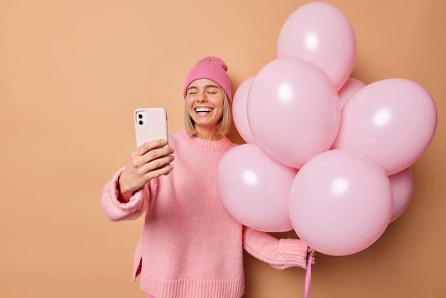 Photo d'une femme blonde heureuse sourit à pleines dents pose pour selfie prend selfie vêtu d'un pull et un chapeau tient un tas de ballons gonflés célèbre une occasion spéciale isolée sur fond marron