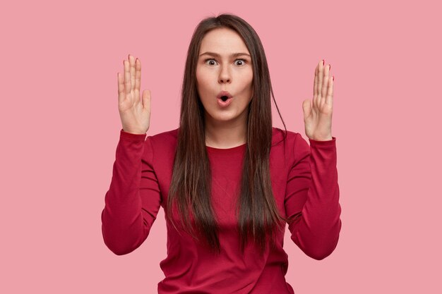 La photo d'une femme brune étonnée montre quelque chose de gros ou d'énorme avec les deux mains, a surpris l'expression du visage, porte une tenue rouge