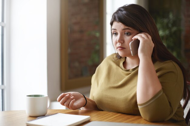 Photo d'une femme caucasienne de taille plus jeune sérieuse parlant au téléphone intelligent assis à la table du café devant un cahier ouvert et une tasse, ayant un regard inquiet, bouleversé par des nouvelles négatives. Mise au point sélective