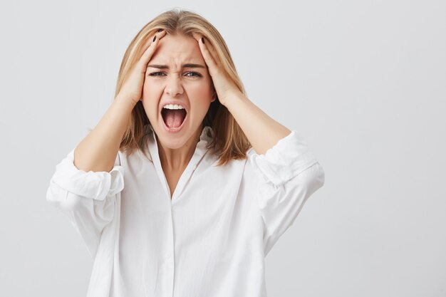 Photo d'une femme déçue aux cheveux blonds tenant ses mains sur les tempes, le visage froncé, la bouche grande ouverte, hurlant de désespoir et de terreur.