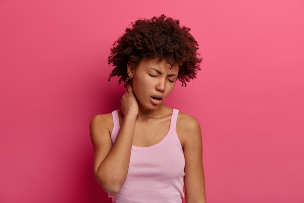 Photo gratuite photo d'une femme en détresse touche le cou, souffre d'une maladie du dos ou de la colonne vertébrale, bâille et ferme les yeux, a une expression fatiguée, habillée avec désinvolture, a des sentiments douloureux, isolée sur un mur rose