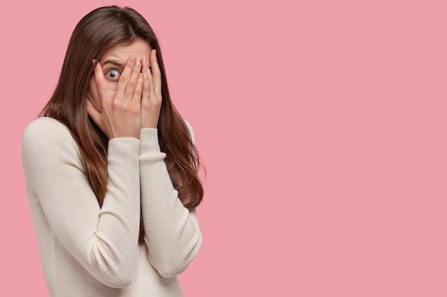 Photo d'une femme effrayée couvre le visage à deux mains, regarde à travers les doigts, regarde avec peur, a les cheveux noirs, vêtue d'un pull confortable