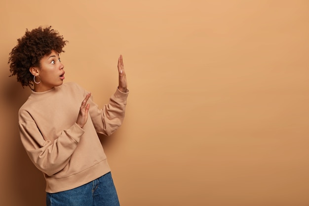 Photo d'une femme émotionnelle effrayée se tourne sur le côté, lève les paumes en signe de défense, regarde avec une expression effrayée, les yeux sont sortis