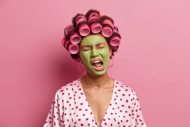 Photo d'une femme ethnique mécontente qui garde la bouche ouverte, ressent de la fatigue, bâille et applique un masque vert, vêtue de vêtements domestiques décontractés, isolée sur rose. Traitement du visage et concept de bien-être