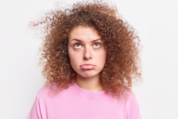 photo d'une femme européenne bouleversée, la lèvre inférieure semble tristement au-dessus d'être déçue par quelque chose qui a des cheveux bouclés et touffus habillés avec désinvolture isolés sur un mur blanc. Expressions du visage.