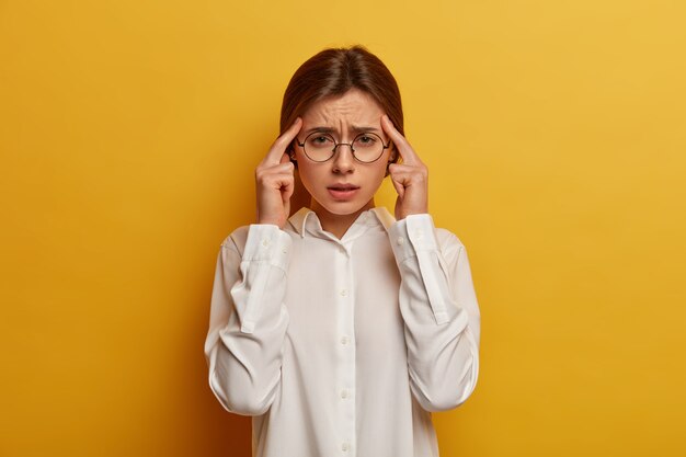 Photo d'une femme européenne épuisée tient les doigts sur les tempes, souffre de maux de tête, réfléchit, sourit narquoisement, habillée d'une chemise blanche élégante, rassemble ses sentiments, réfléchit à une décision importante