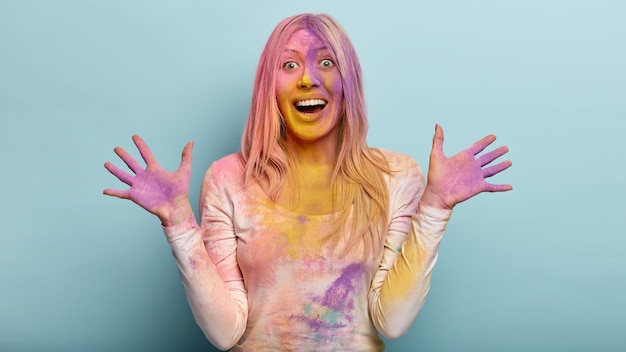 Photo d'une femme européenne heureuse montre des paumes tachées de violet, s'étire les mains, rit joyeusement, fait des gestes avec émotion, modèles contre le mur bleu, s'amuse sur l'événement festif annuel de Holi en Inde